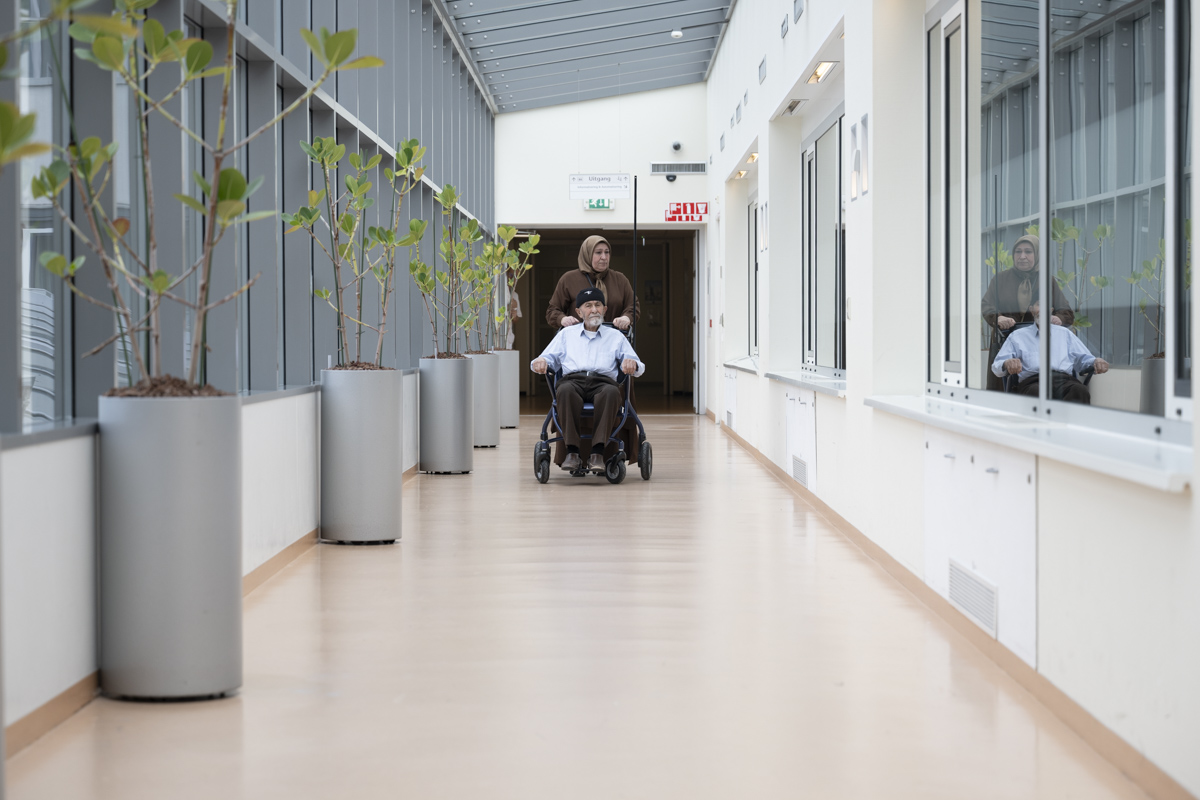 Naaste duwt patiënt in rolstoel door lange gang 